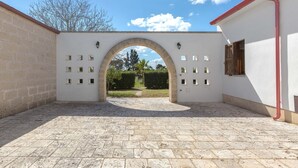 Un patio al aire libre con suelo de piedra, una pared blanca con pequeñas ventanas y una puerta de madera.