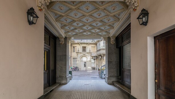 A corridor with ornate ceiling and large doors leading to a building with balconies.