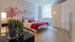 A modern bedroom with a bed, a nightstand, a potted plant, and a window with sheer curtains.