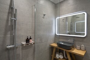 A modern bathroom with a glass-enclosed shower, a wooden vanity with a sink, and a mirror with integrated lighting. - AKRI Suites Vourvoulos (Santorini)