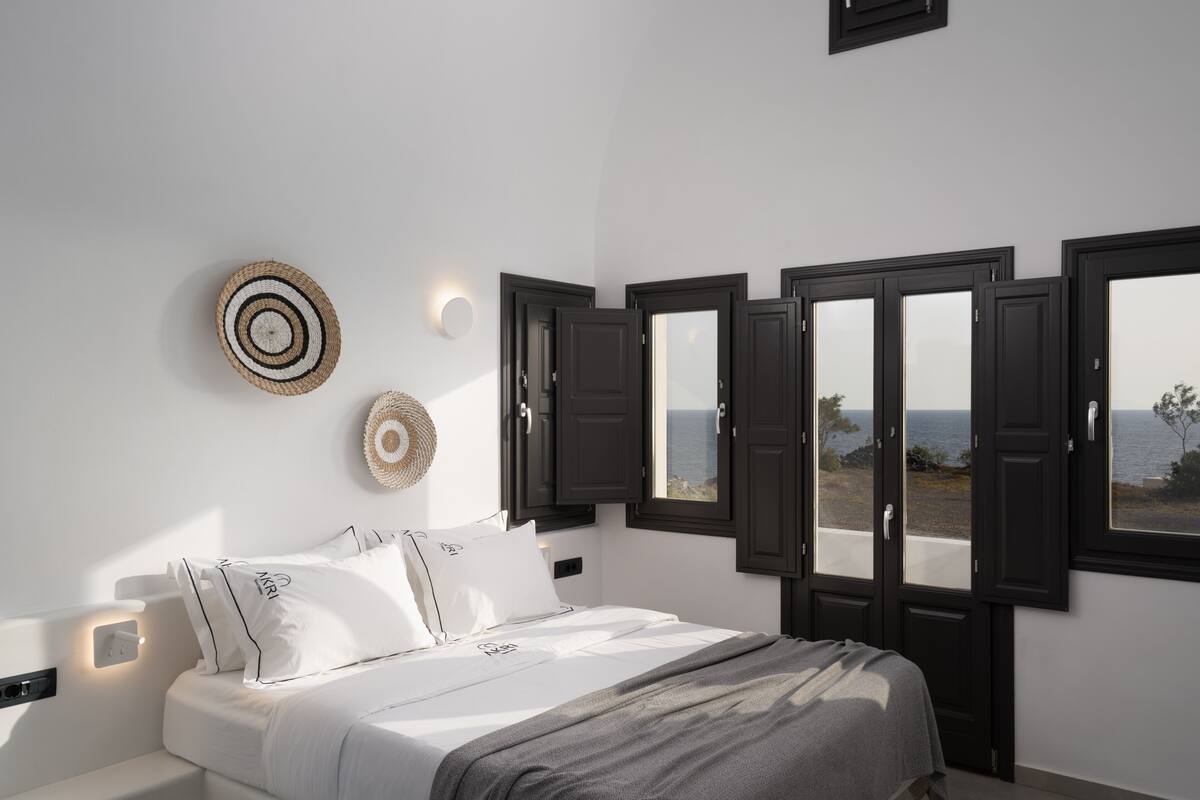 Quarto com cama, roupa de cama branca, vista para o mar através de janelas abertas e luminária fixada na parede.