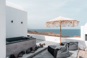 A modern outdoor seating area with a hot tub, a large woven umbrella, and a view of the sea. - AKRI Suites Vourvoulos (Santorini)