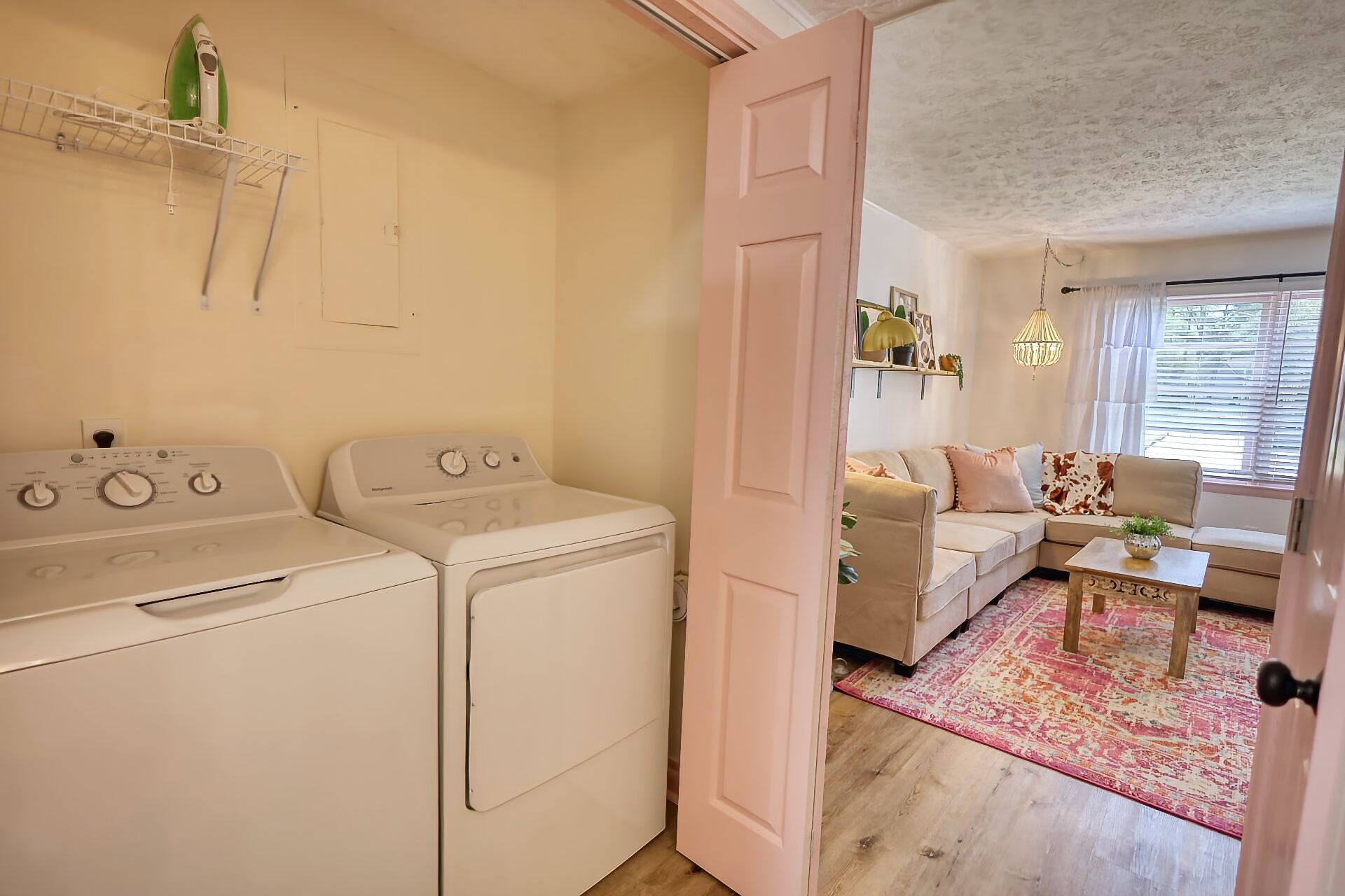 Une buanderie équipée de deux sèche-linge blancs, d’un lave-linge et d’une porte rose donnant sur un espace de vie comprenant un canapé, une table basse et une fenêtre avec des stores.