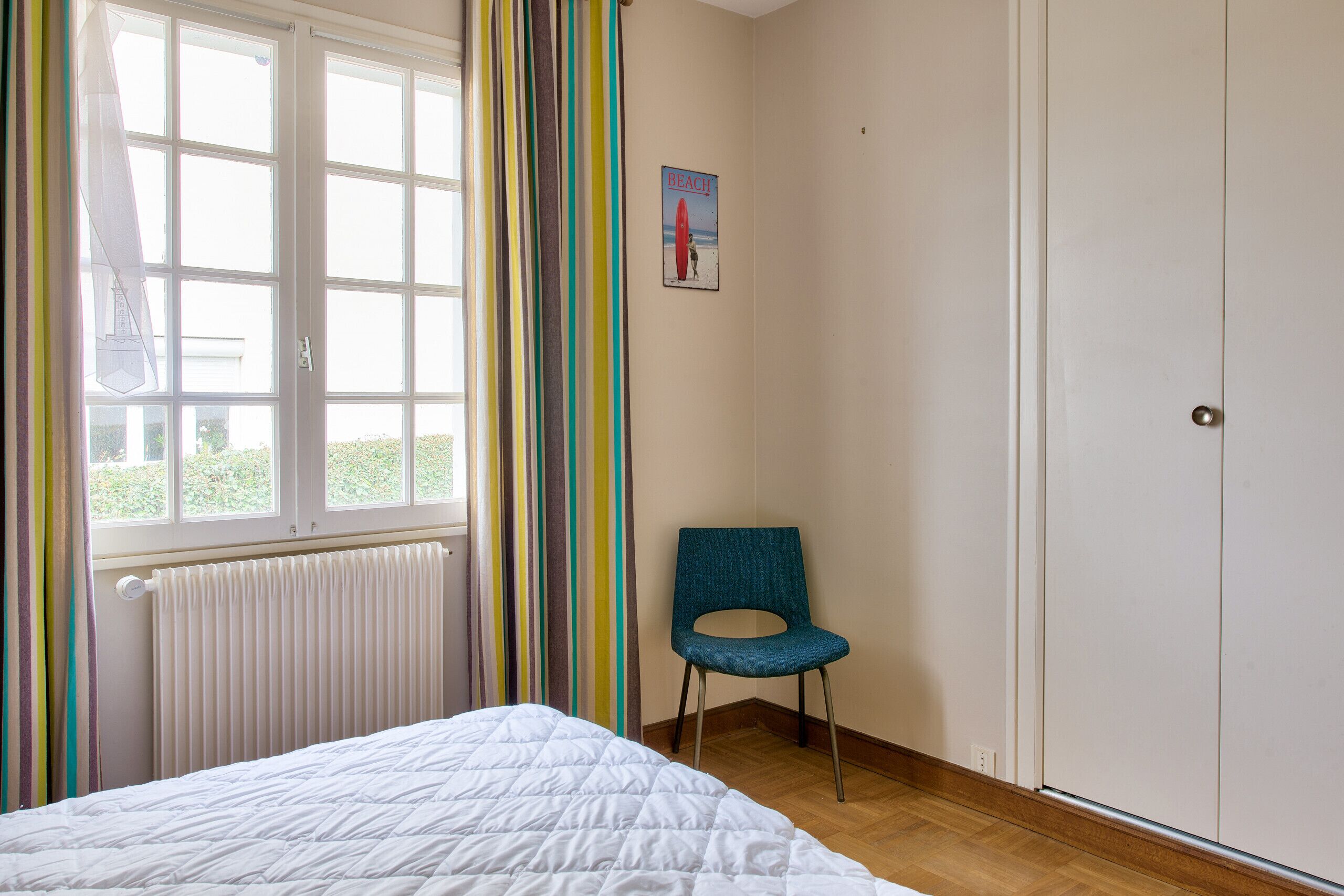 Une chambre à coucher avec un lit, une chaise, un radiateur, une fenêtre avec des rideaux et une porte.