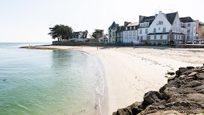 A beach with a row of white and blue buildings, a clear sky, and calm sea. - 800 m from harbour – House terrace BBQ – Sleeps 12 (Loctudy)