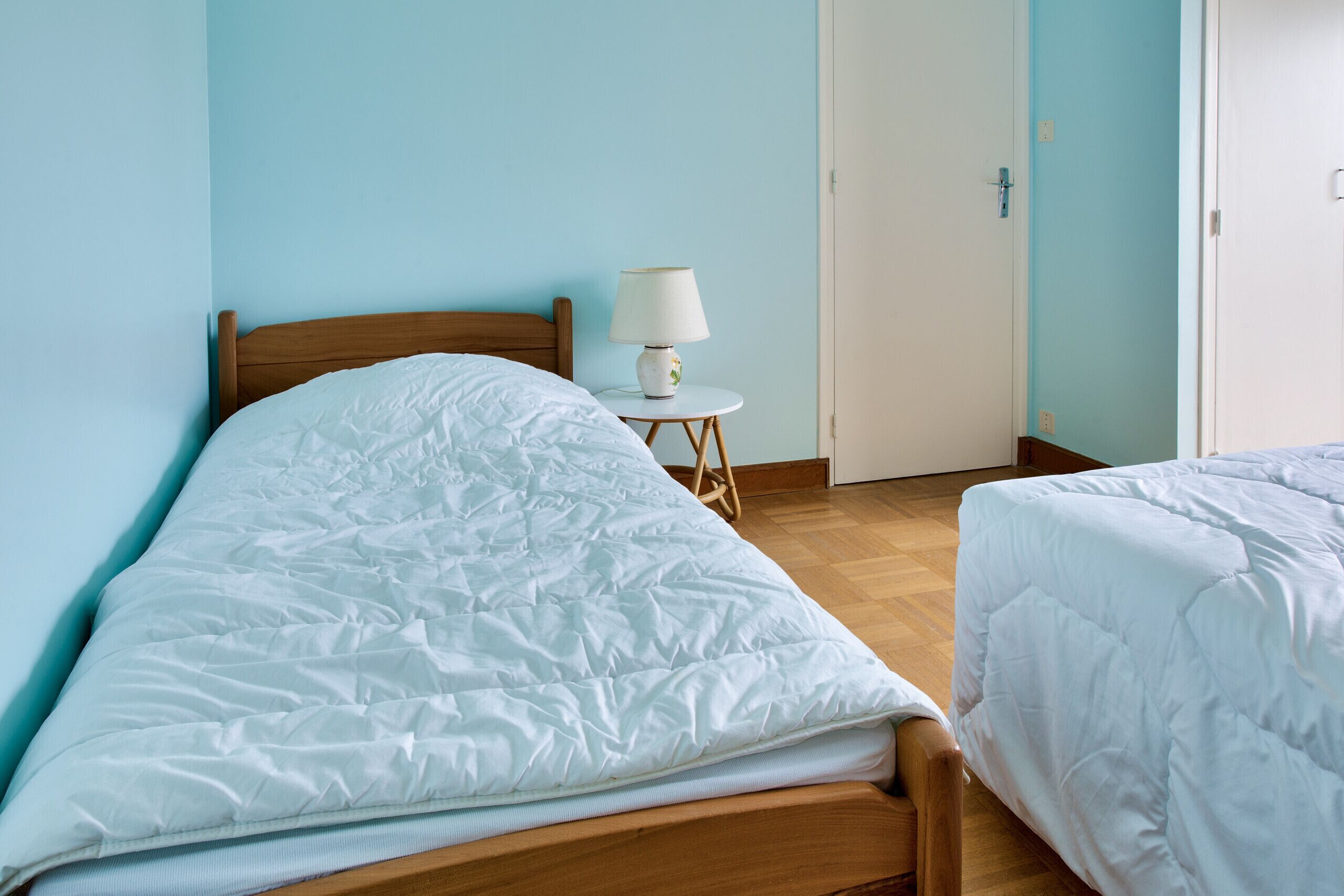 A bedroom with a wooden bed, a white bedside table with a lamp, and a door in the background.