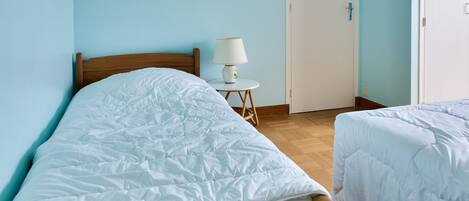 A bedroom with a wooden bed, a white bedside table with a lamp, and a door in the background.