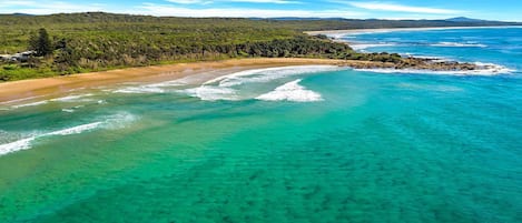 A coastal landscape with clear turquoise waters, sandy beach, and a forested area.