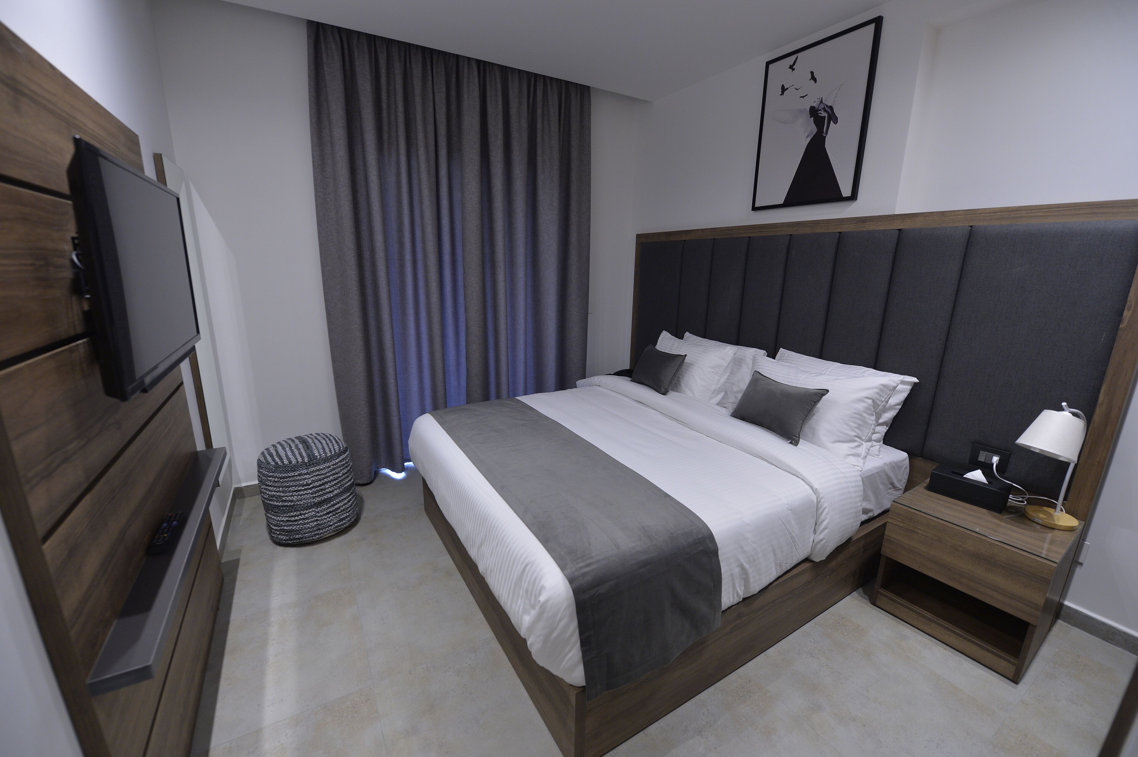 A modern hotel room with a large bed, a flat-screen TV, a bedside table with a lamp, and a large window with curtains.