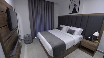 Quarto de hotel moderno com uma cama grande, televisão de tela plana, mesa de cabeceira com luminária e uma janela ampla com cortinas.