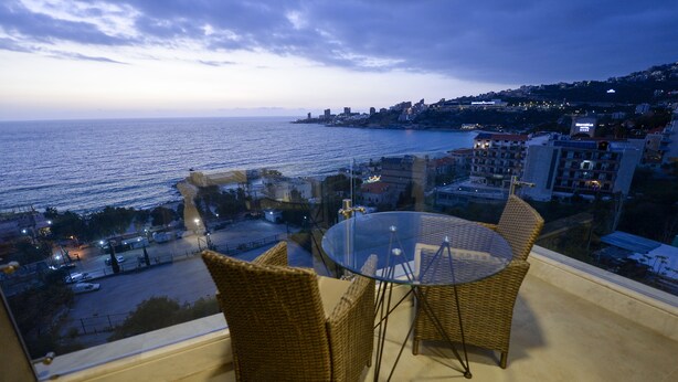 A balcony with wicker chairs and a glass table overlooking a coastal cityscape at dusk. - ZETT HOTEL (Jounieh)