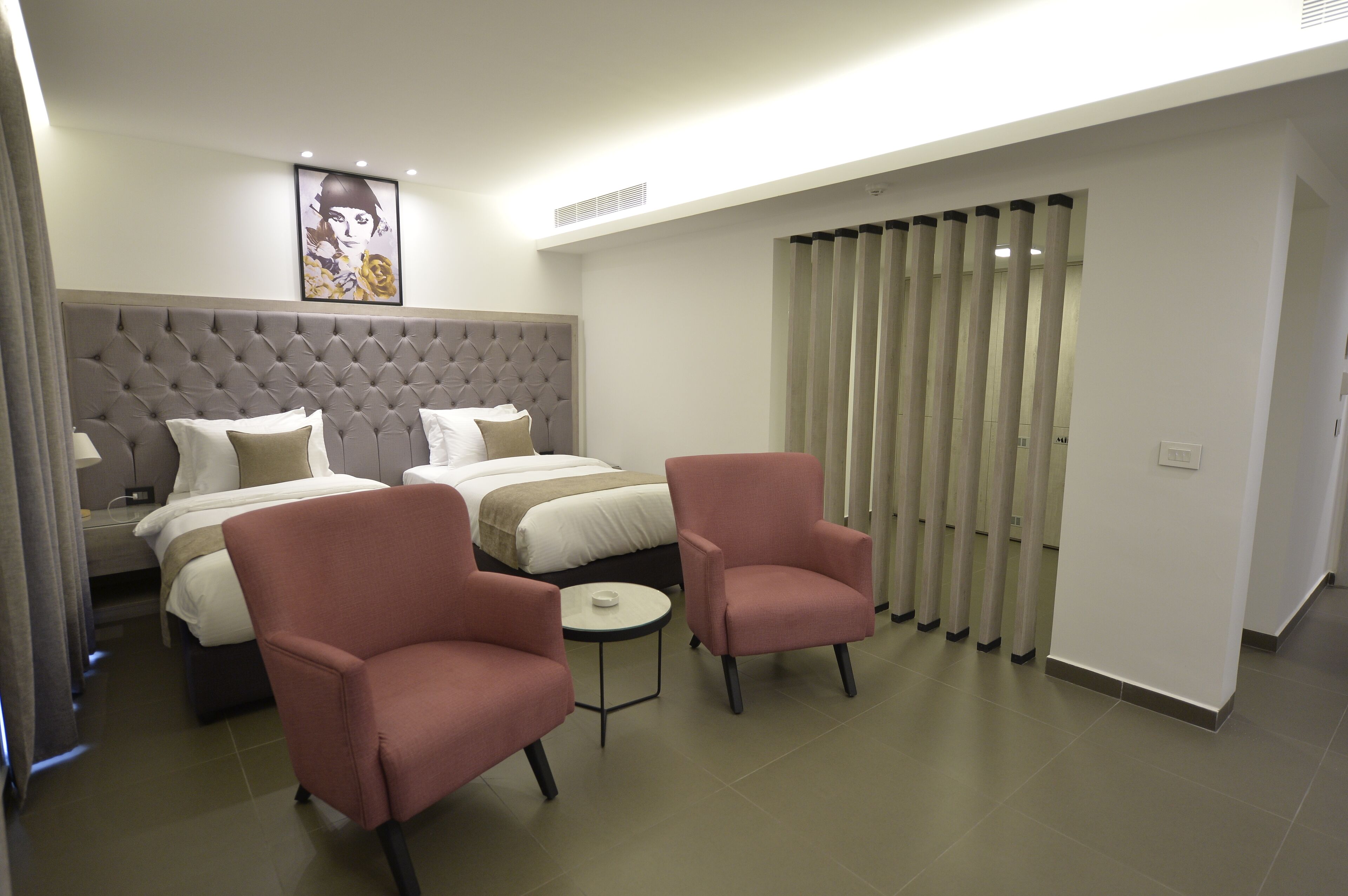 Habitació d'hotel amb dos llits, un panell de capçalera gris amb textura, una taula rodona petita i dues butaques roses.