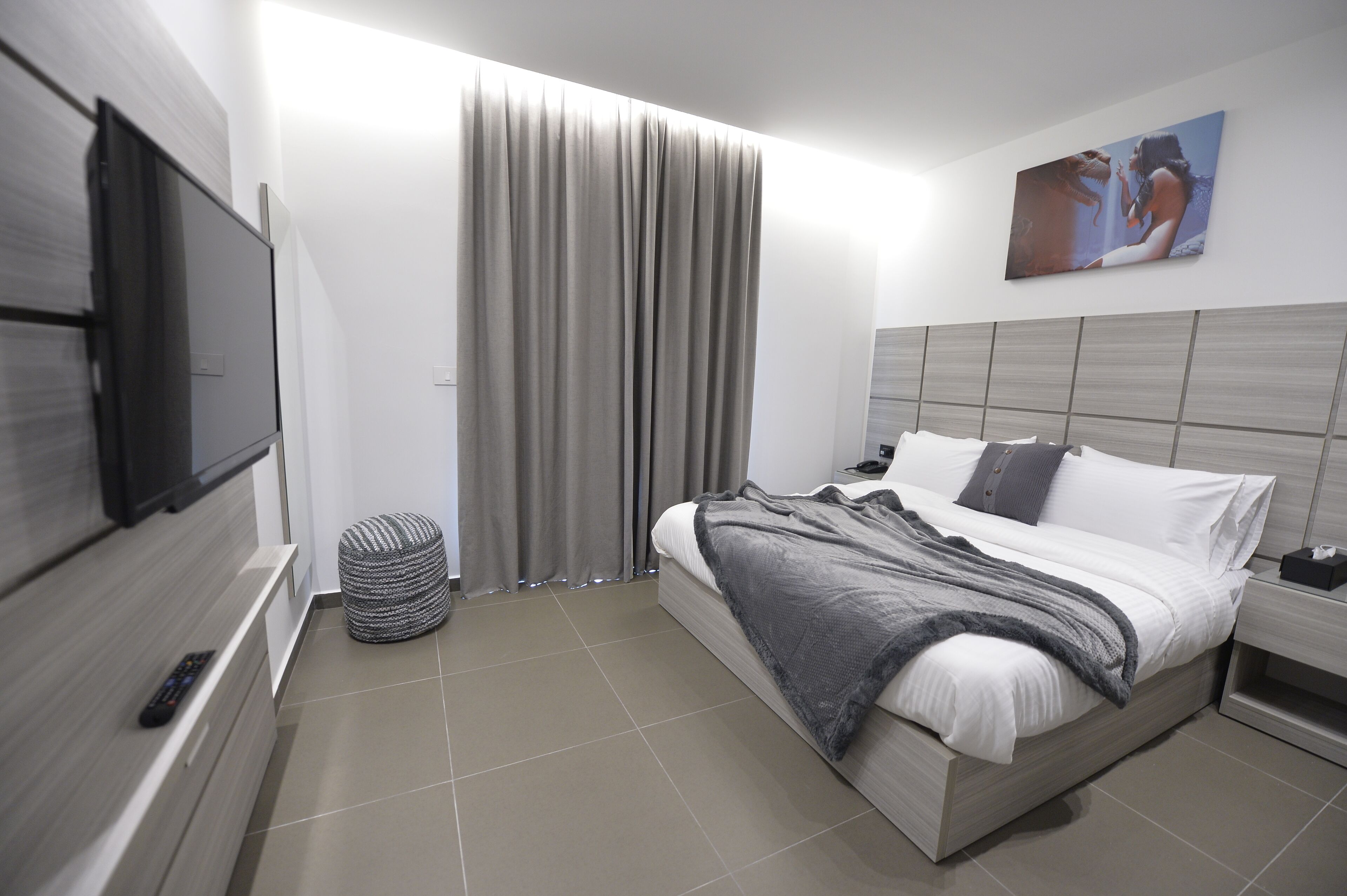 A modern hotel room with a bed, a TV mounted on the wall, a curtain, and a painting on the wall.