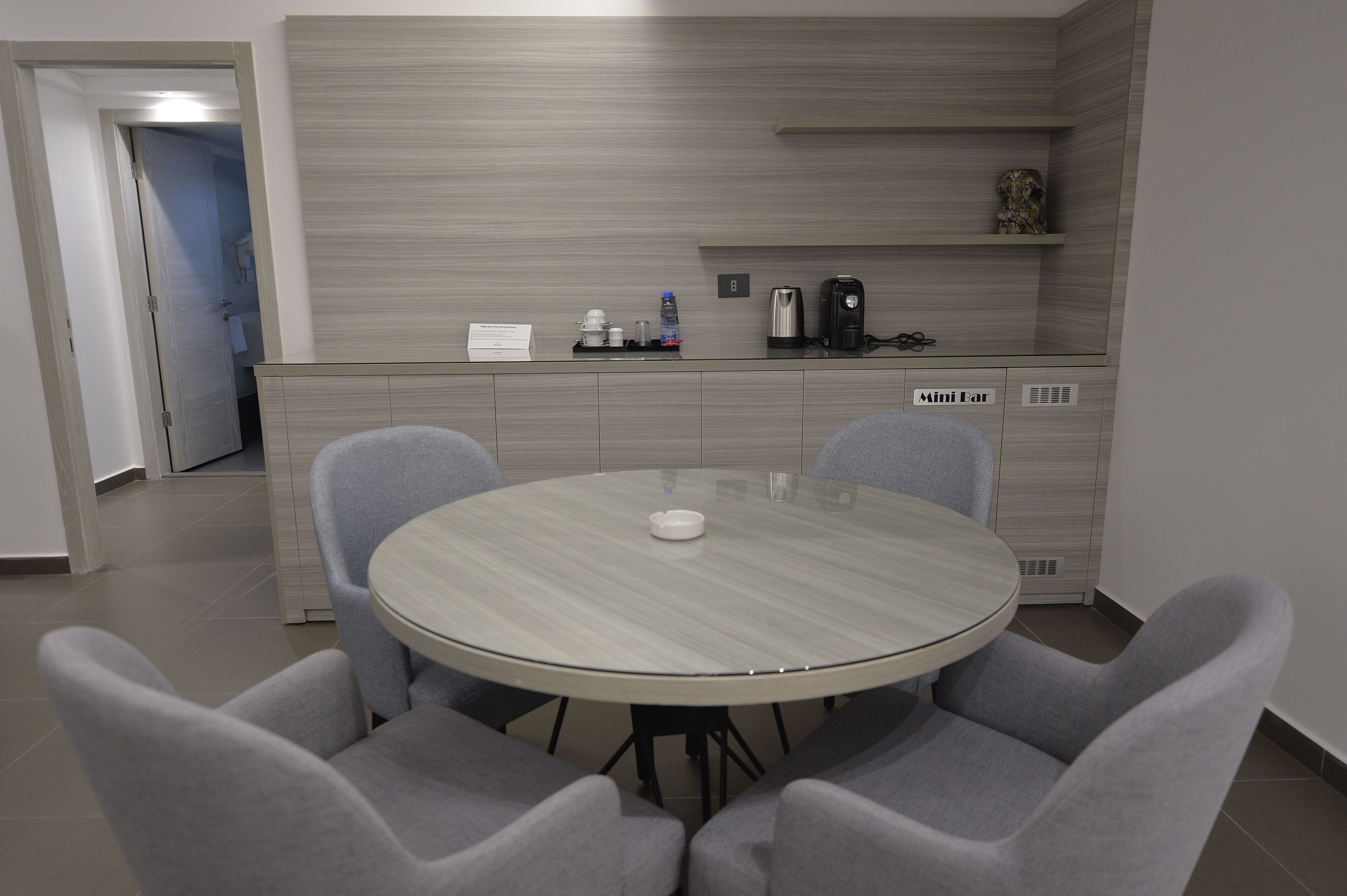 A modern kitchen with a round table and grey chairs, a coffee machine, and a mini fridge.