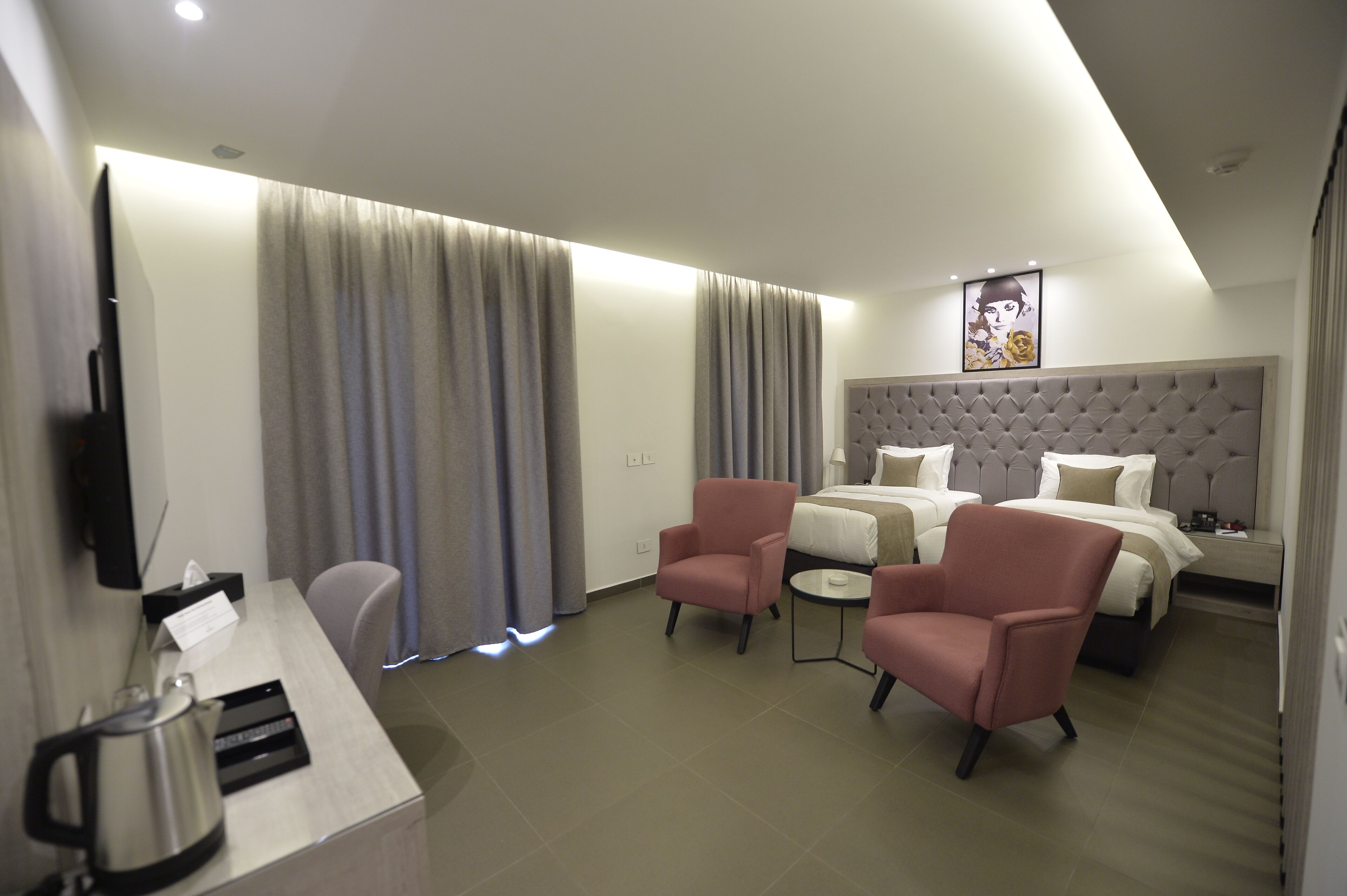 Habitación de hotel con una cama, dos sillones rosados, una mesita, un televisor y una cafetera.