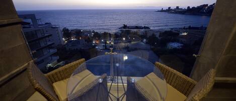 A balcony with a glass table and wicker chairs overlooking a coastal cityscape at dusk.