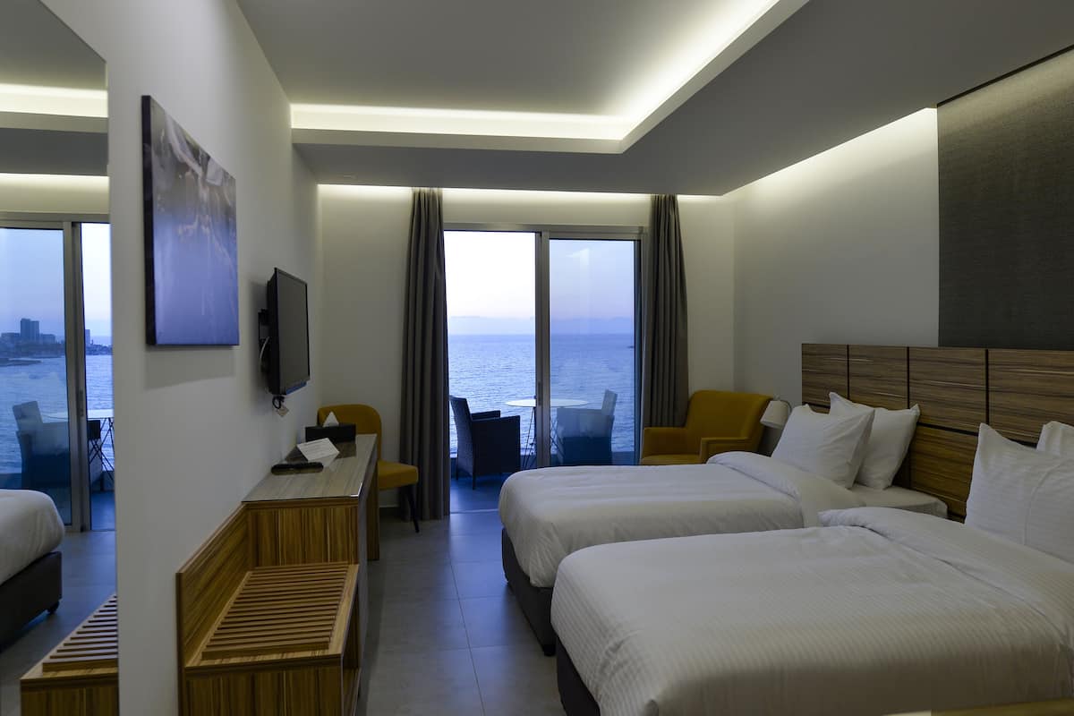 Habitación de hotel con dos camas, un escritorio, una silla y un televisor. Se observa un ventanal con vistas al mar.