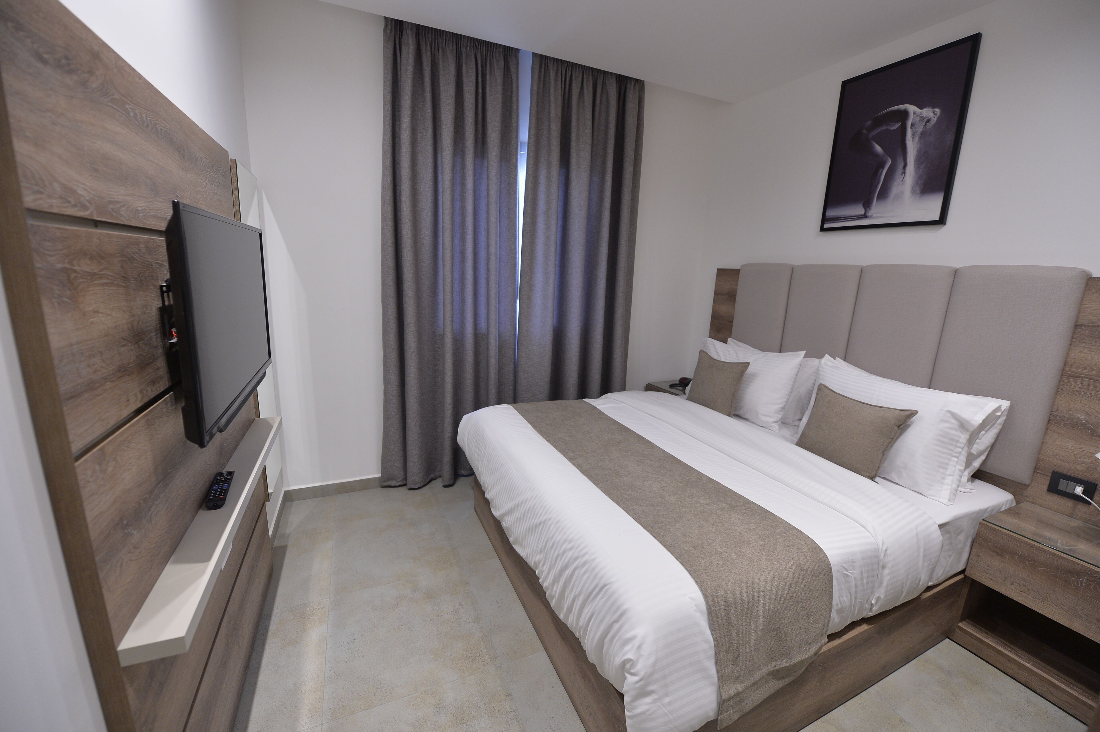 Quarto de hotel moderno com uma cama grande, uma televisão de tela plana fixada em um painel de madeira, uma mesinha de cabeceira e uma janela com cortinas.
