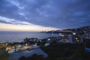 Paisagem urbana costeira ao entardecer, com edifícios à beira-mar, uma estrada que leva à praia e uma vista clara do mar.