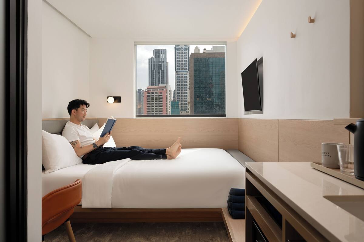 A modern hotel room with a bed, a small desk, a window with a city view, and a flat-screen TV mounted on the wall.