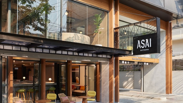 Un bâtiment moderne à la façade vitrée, sur lequel on peut lire l’inscription « ASAI SATHORN », et un espace extérieur aménagé avec des chaises et des tables.