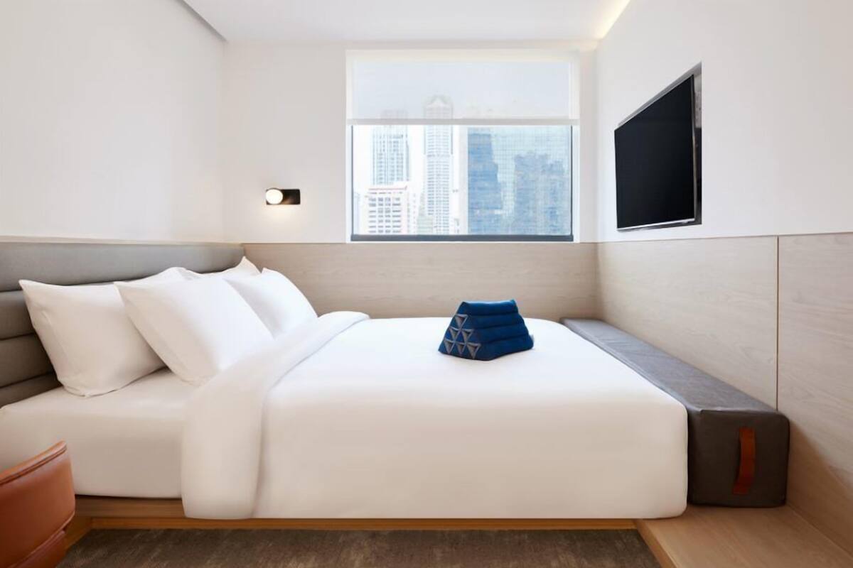 A modern hotel room with a bed, a seating area, a TV, and a window with a city view.