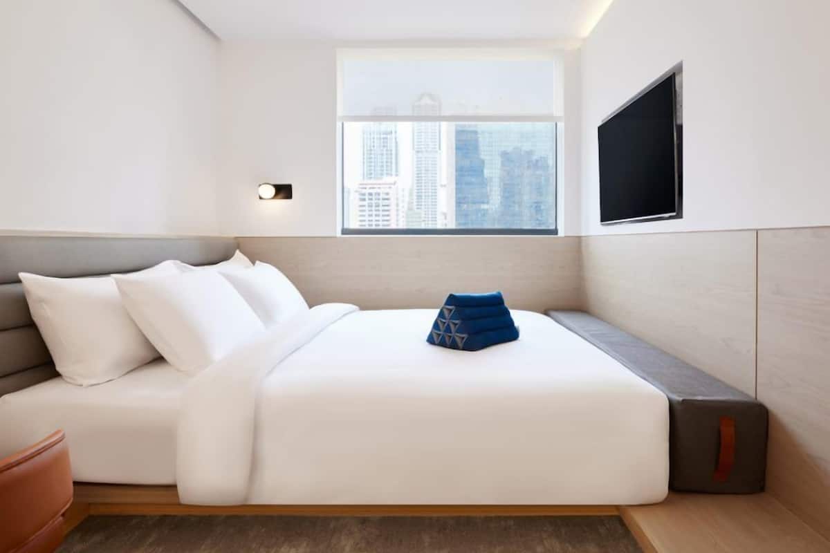 A modern hotel room with a bed, a seating area, a TV, and a window with a city view.