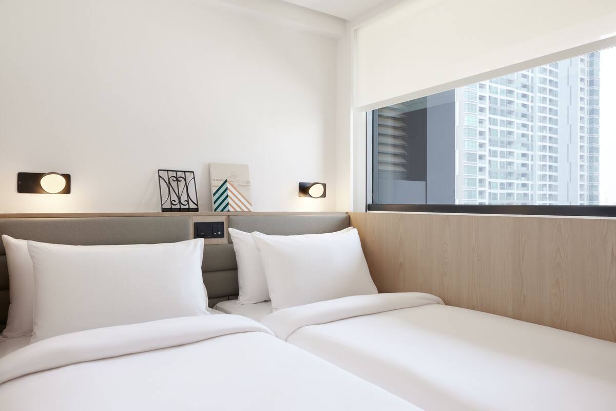 A hotel room with two beds, a wooden headboard, wall-mounted lights, and a large window with a city view.