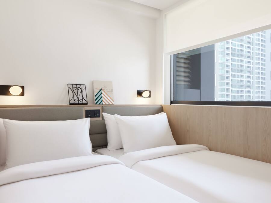 A hotel room with two beds, a wooden headboard, wall-mounted lights, and a large window with a city view.