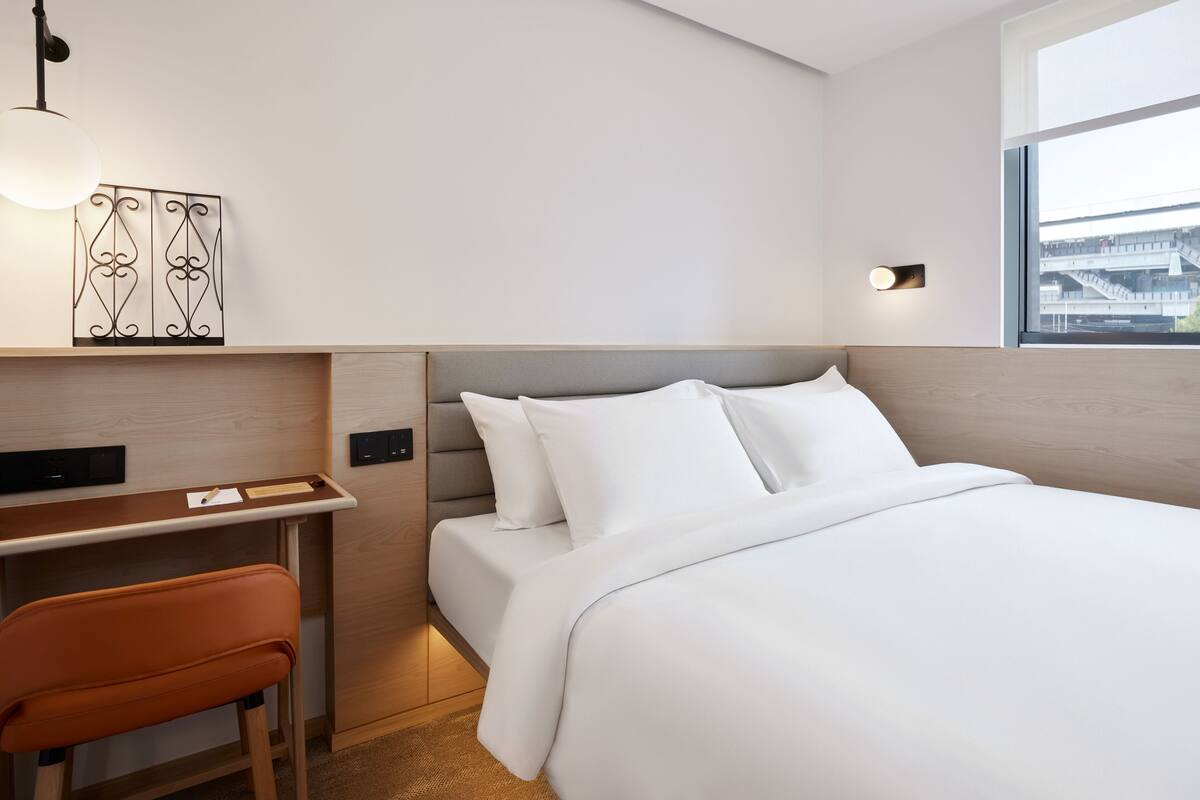A hotel room with a bed, desk, chair, and a view of a building through the window.