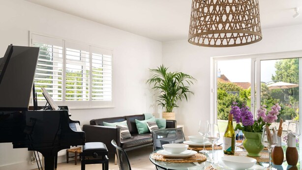 Un salon comprenant un grand piano, un canapé, une table à manger dressée pour quatre personnes et une grande fenêtre avec des stores.