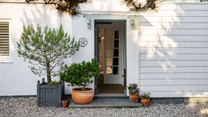 Un bâtiment blanc avec une porte sur laquelle on peut lire « Bienvenue » et « Studio 17 ». Devant, on trouve des plantes en pot et un petit arbre.