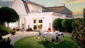 Une maison à deux étages dotée d’un patio comprenant une table et des chaises, le tout entouré d’une pelouse bien entretenue et de haies.