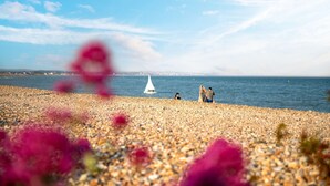 Une plage de galets, avec un voilier à l’horizon, un ciel dégagé et quelques personnes sur la rive.