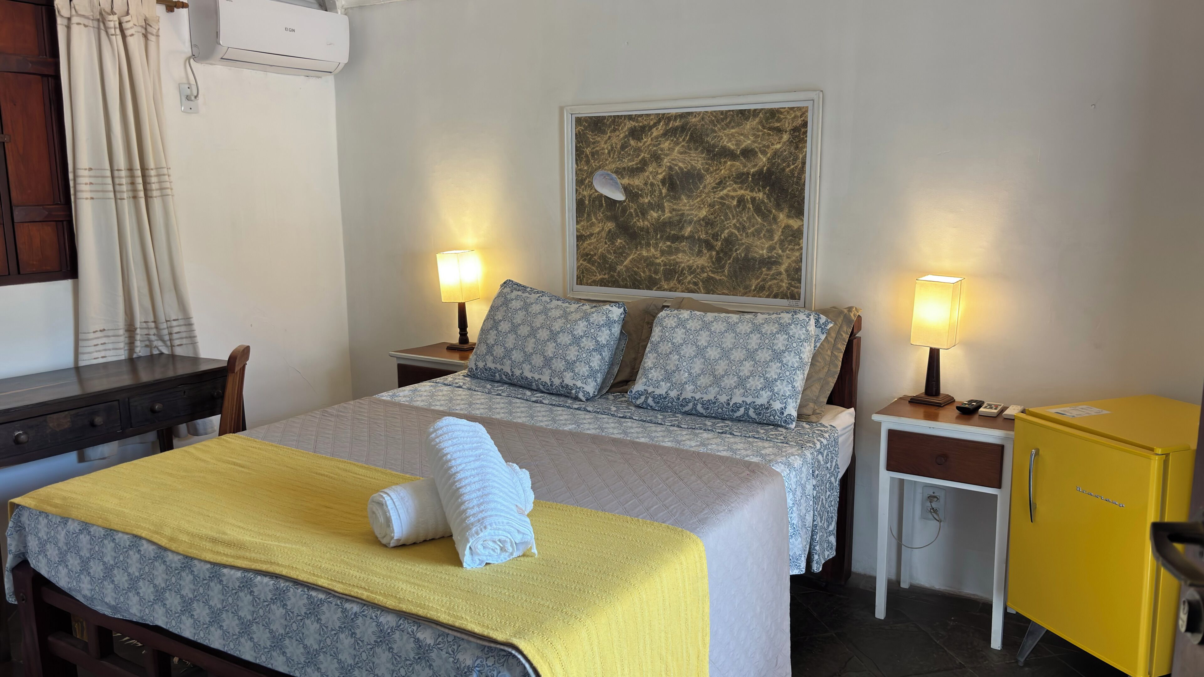 Una cama bien tendida con ropa de cama estampada, dos lámparas de noche, un pequeño armario amarillo y un cuadro en la pared.