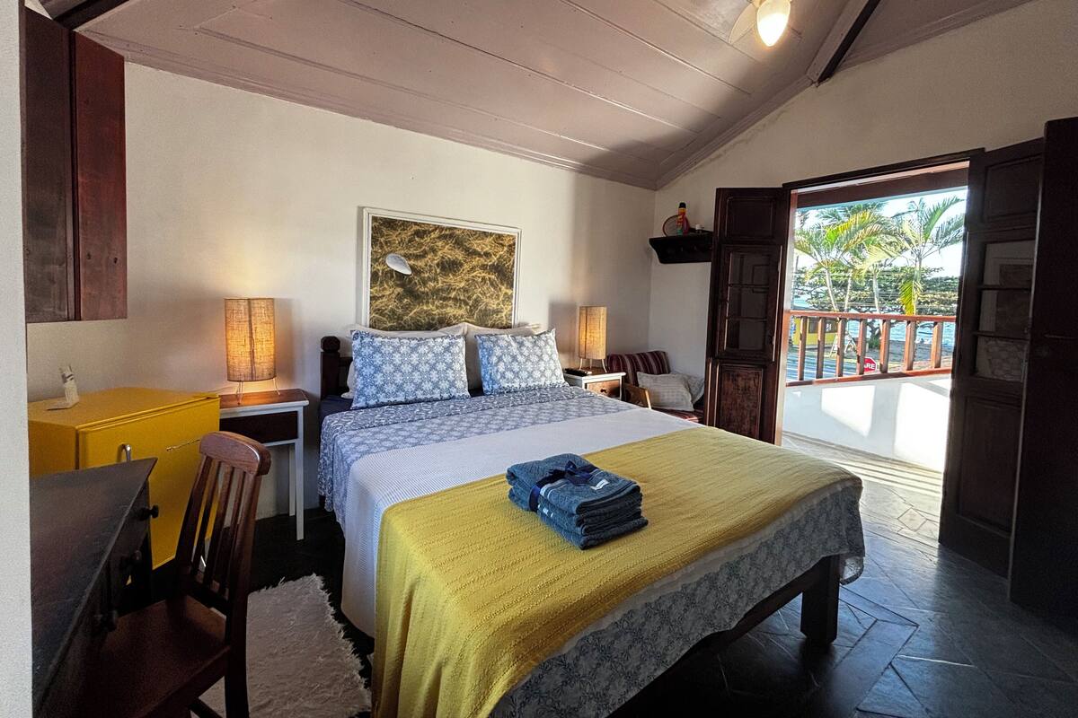 Un dormitorio con cama, mesitas de noche, ventilador de techo, ventana con vista a palmeras y puerta que da al exterior.