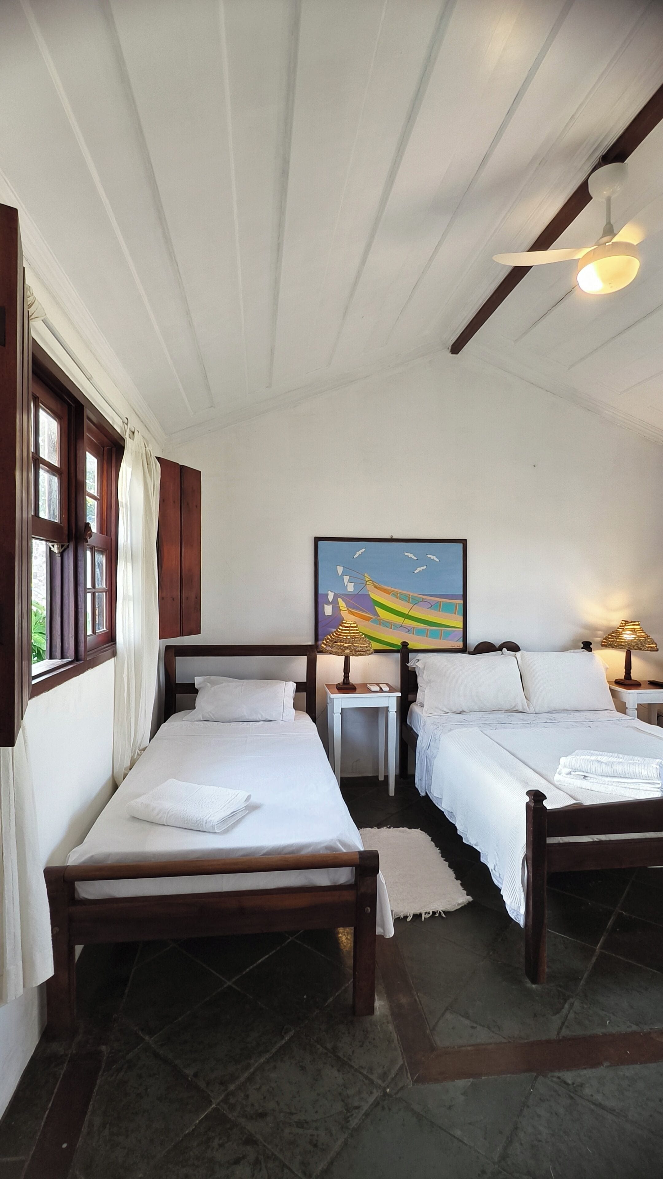 A bedroom with two single beds, a painting on the wall, a ceiling fan, and a window with curtains.