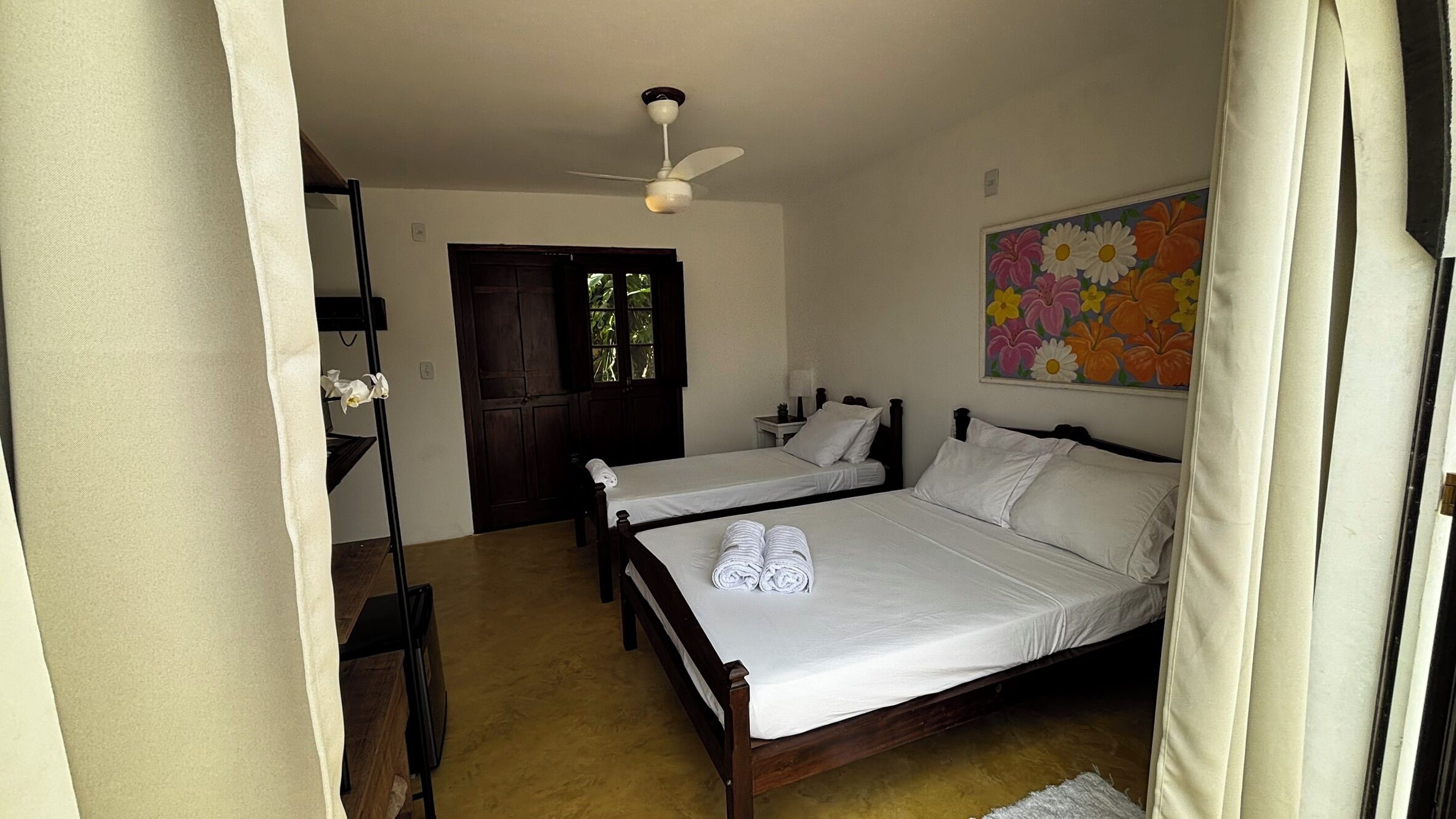 Quarto com duas camas, um ventilador de teto, uma porta e uma pintura floral na parede.