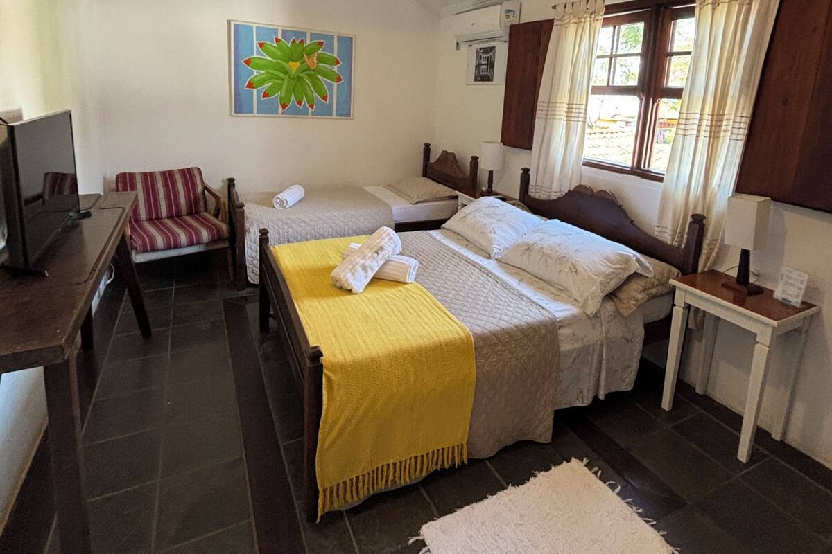 Una habitación con cama, una silla, una mesa, televisión, ventilador de techo y ventana con cortinas.