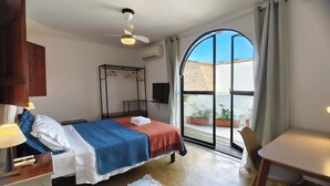 Un dormitorio con cama, escritorio, silla, armario, ventilador de techo, televisor y un ventanal con vistas al exterior.