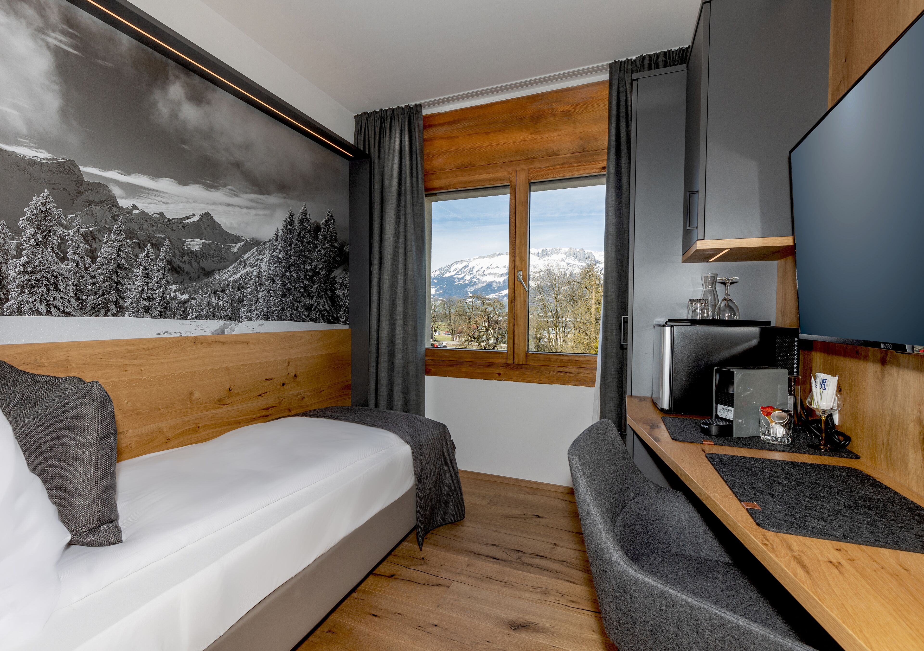 A modern hotel room with a bed, a seating area, a TV, and a window with a mountain view.