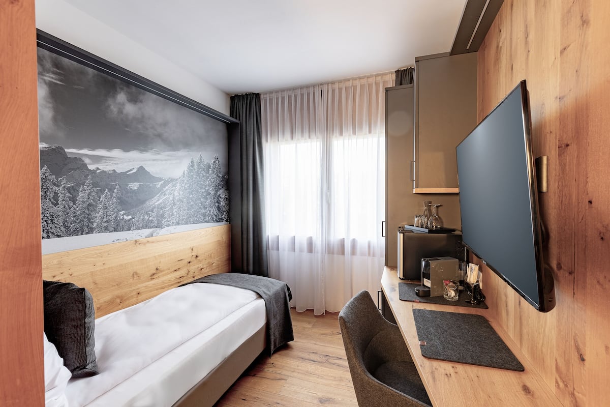 Une chambre d’hôtel moderne équipée d’un lit, d’un bureau, d’une télévision à écran plat et d’une grande fenêtre avec des rideaux légers.