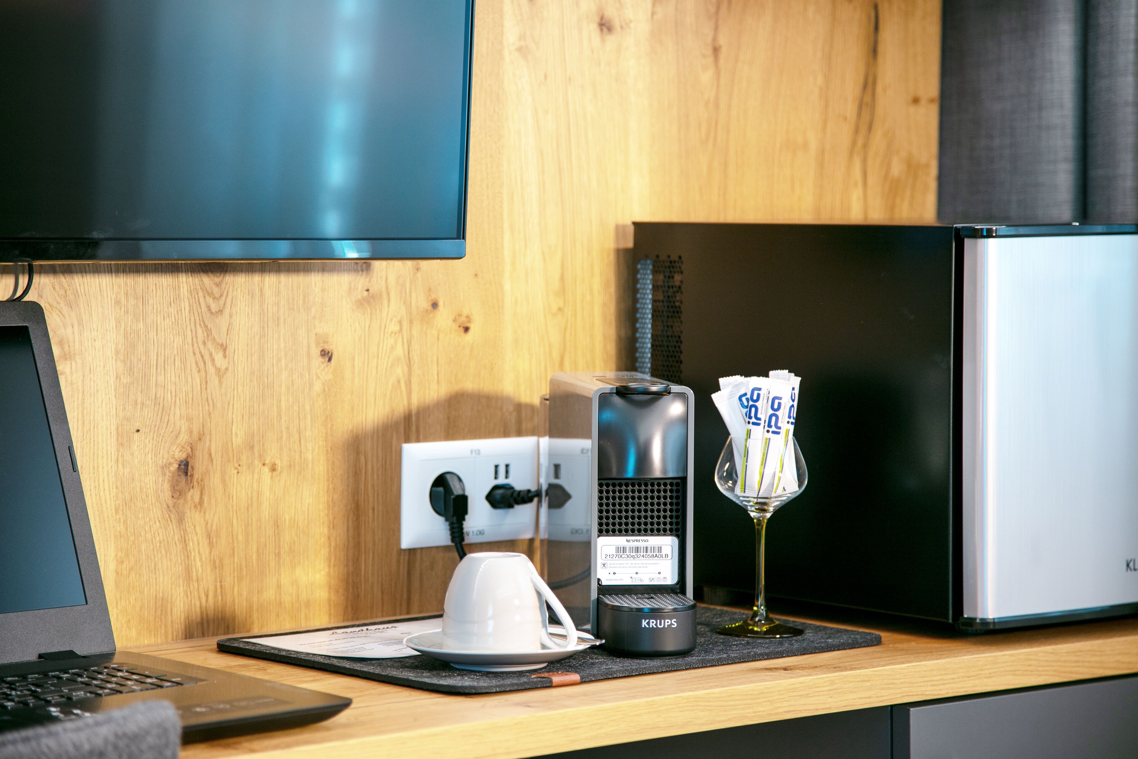 Un bureau en bois avec un ordinateur portable, une machine à café Krups, une tasse de café et un réfrigérateur.