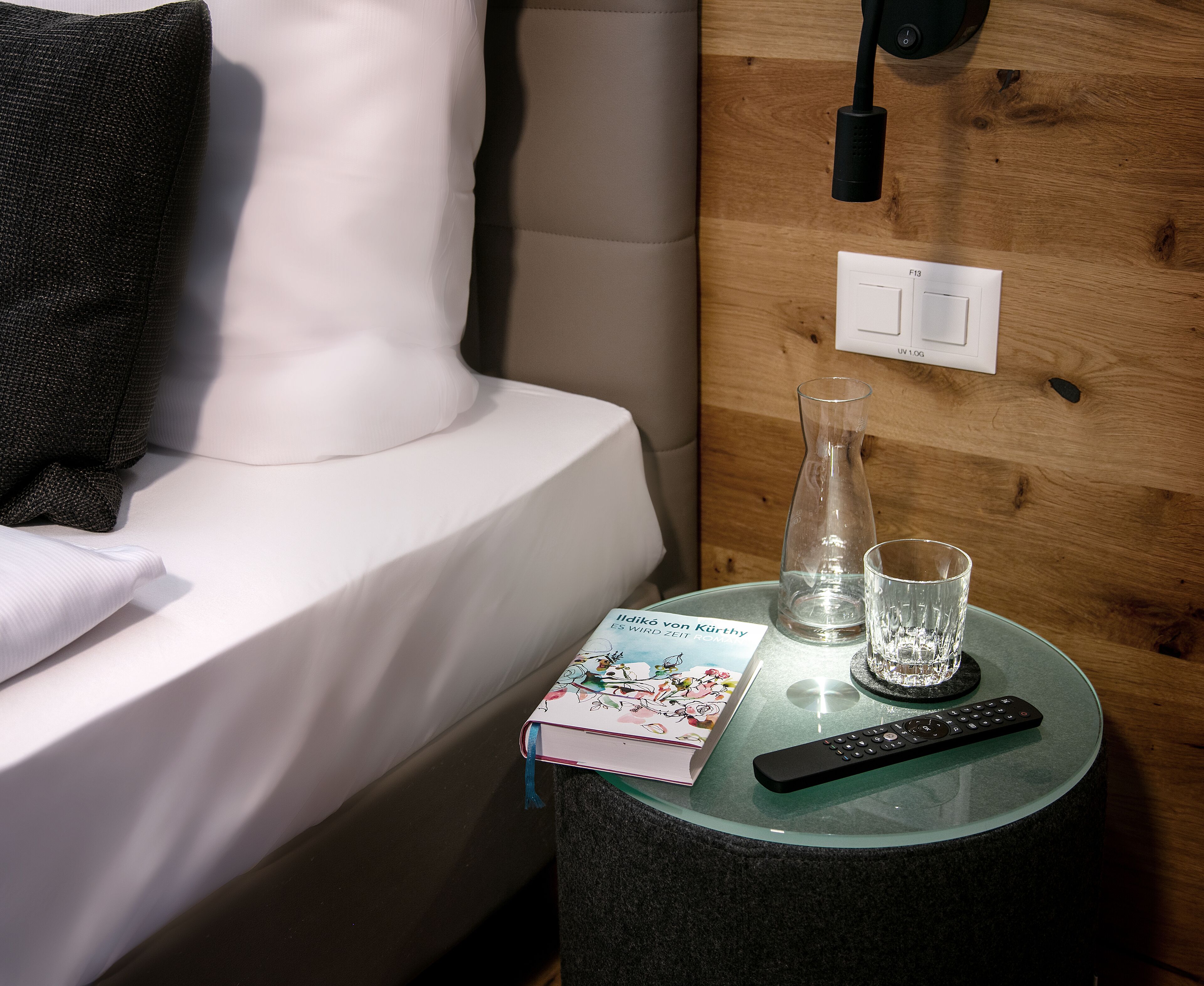 Un lit bien fait, avec un oreiller, une table de chevet sur laquelle se trouvent un livre, un verre et une télécommande, ainsi qu'une prise électrique au mur.