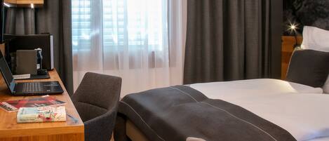 A hotel room with a bed, a desk with a laptop, a chair, and a red suitcase.