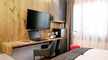 Ein Hotelzimmer mit einem Holztisch, einem Laptop, einem Fernseher, einem roten Koffer und einem Bett.