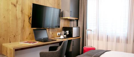 A hotel room with a wooden desk, a laptop, a television, a red suitcase, and a bed.