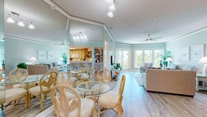 Un salón espacioso con una mesa de comedor de vidrio, sillas de mimbre, un sofá color beige y una cocina al fondo.