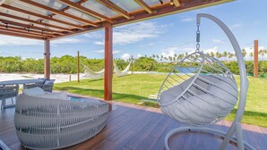 Un espacio de estar al aire libre cubierto con una silla colgante y un banco acolchado, con vistas a un paisaje cubierto de césped y palmeras.