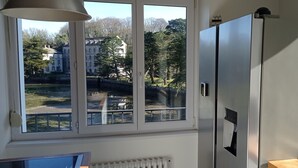 A kitchen with a large window, a refrigerator, and a view of a park and buildings. - Waterfront villa with panoramic sea view (Logonna-Daoulas)